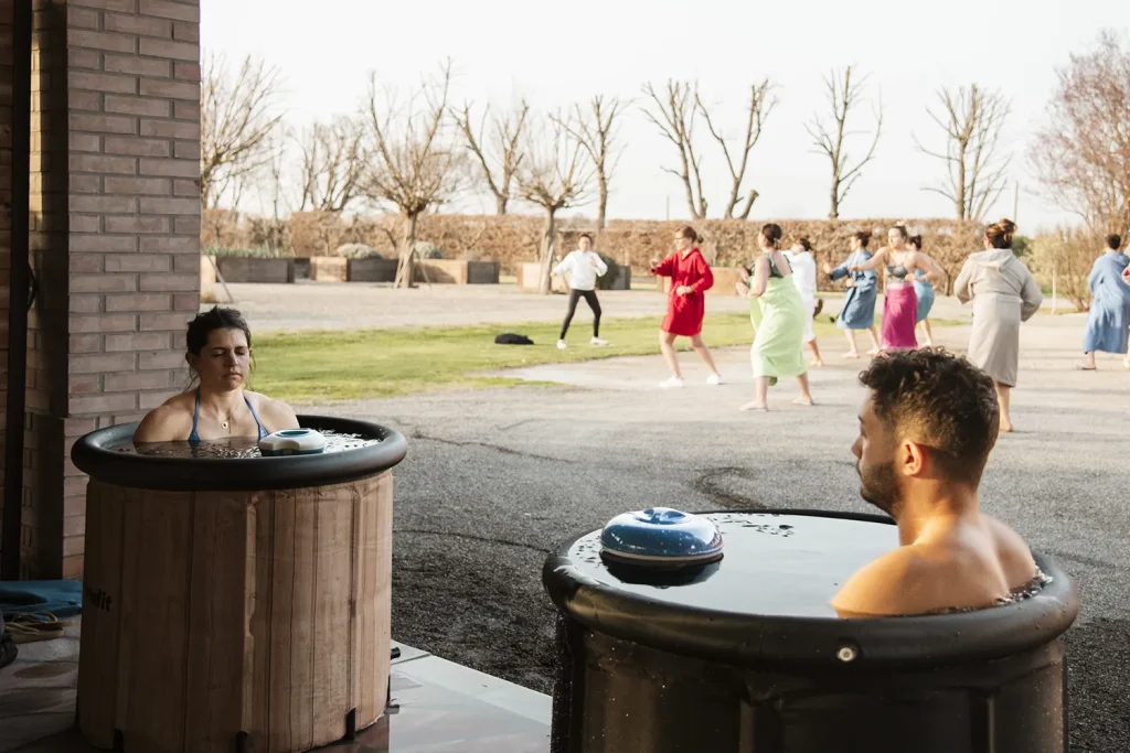 Due persone sono immerse in vasche di legno per l'ice bath mentre sullo sfondo i loro compagni fanno tai chi in un parco.