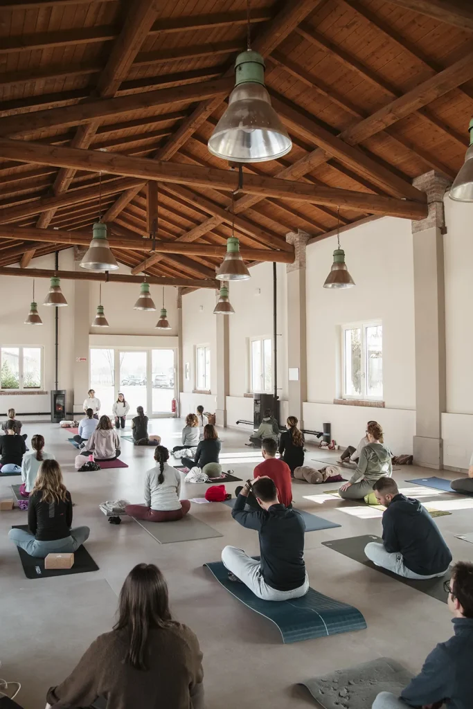 Lezione dii yoga in ampia aula con soffitti in rìtravi di legno e numerose finestre piene di luce.